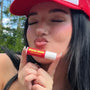 Woman wearing a red cap puckering lips while holding Space Camp Juicy Watermelon lip balm outdoors.