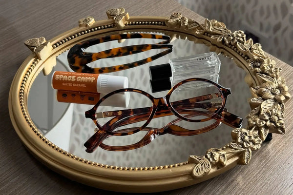 Clean lip balm labeled “Space Camp Salted Caramel” placed on an ornate mirror with tortoiseshell glasses and accessories.