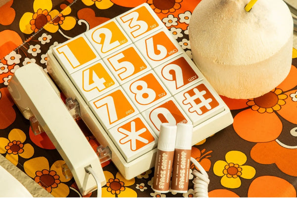Coconut lip balm tubes labeled “Space Camp Coconut Cream” placed next to a retro phone and coconut on a floral tablecloth.