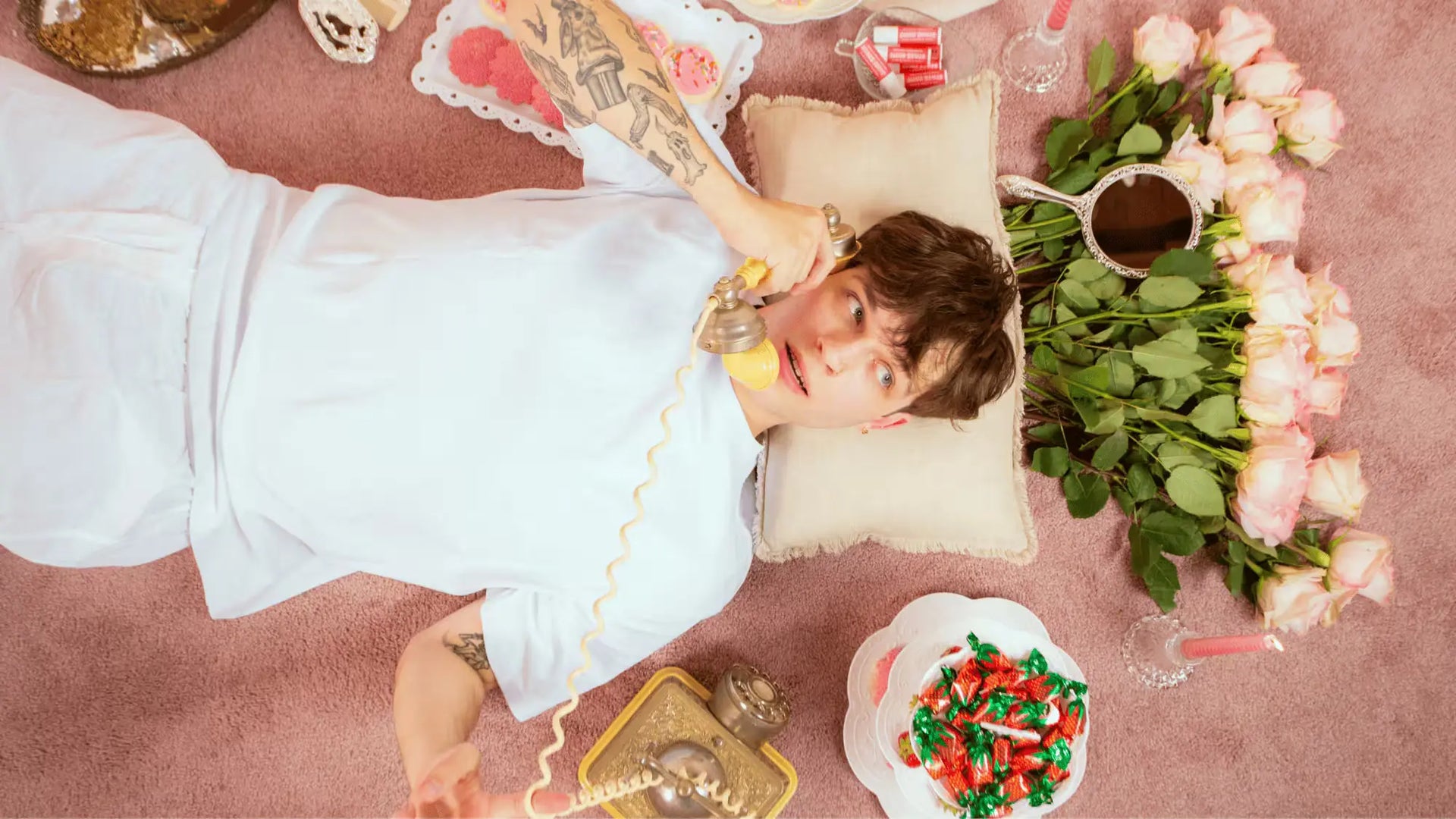  Young man lying on a pink carpet, talking on a vintage phone surrounded by roses and candy dishes