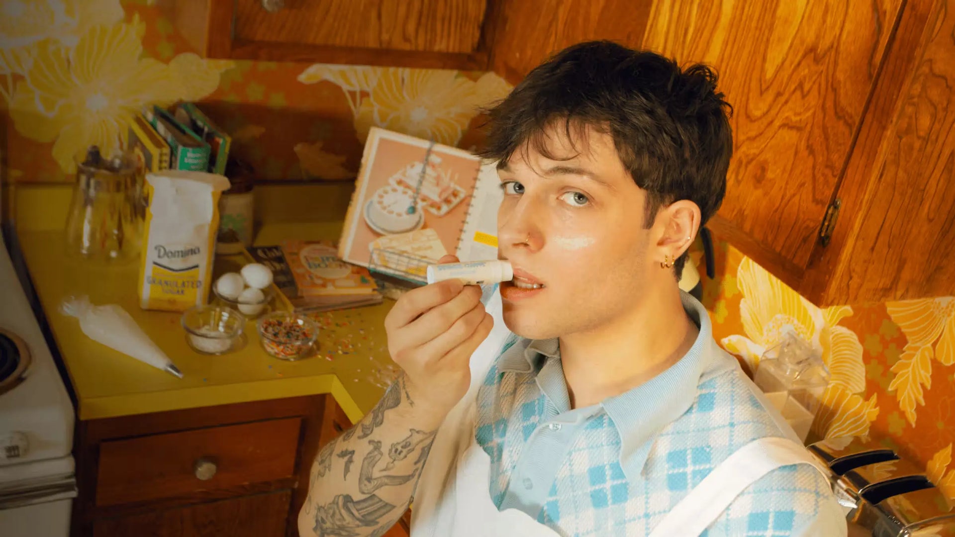  Man applying lip balm in a retro kitchen, surrounded by baking supplies & cookbooks on a yellow countertop