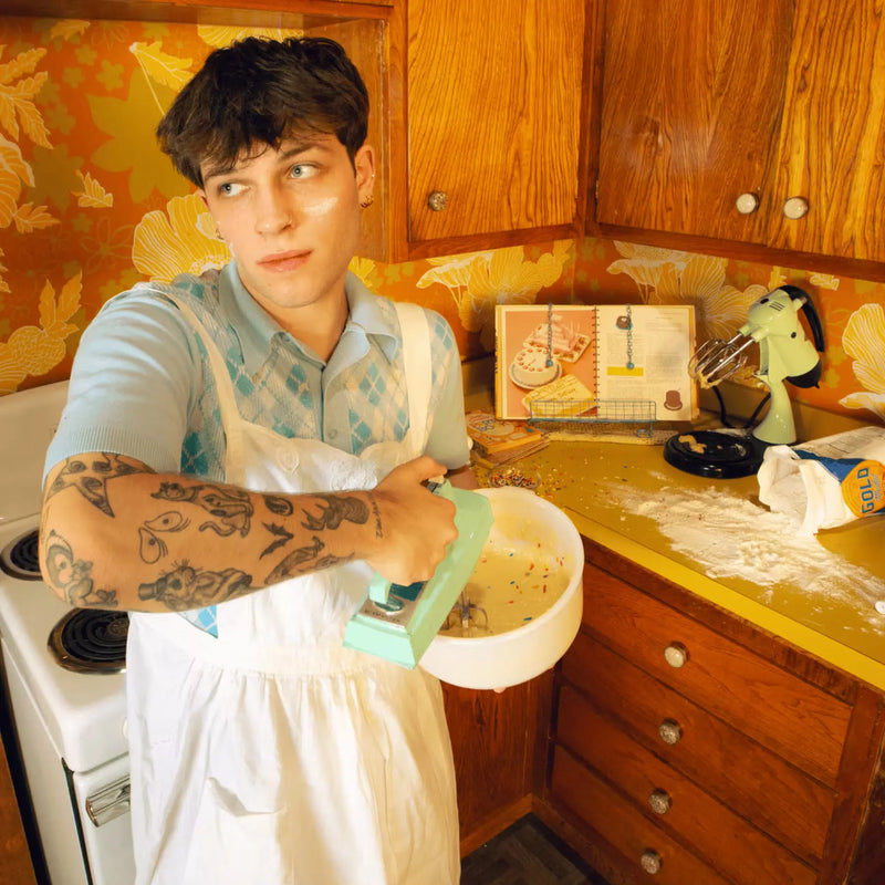 Man in a retro kitchen wearing an apron, holding a bowl with a hand mixer, surrounded by baking ingredients & an open cookbook