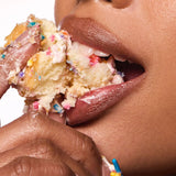 A close-up of a person biting into a slice of birthday cake with colorful sprinkles and frosting.