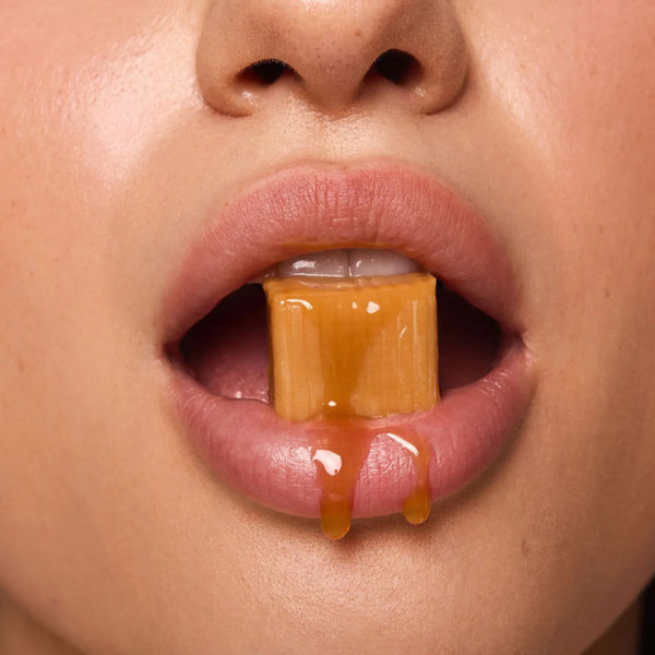 A close-up of a person's lips holding a melting caramel candy that drips down the chin, evoking the salted caramel flavor.
