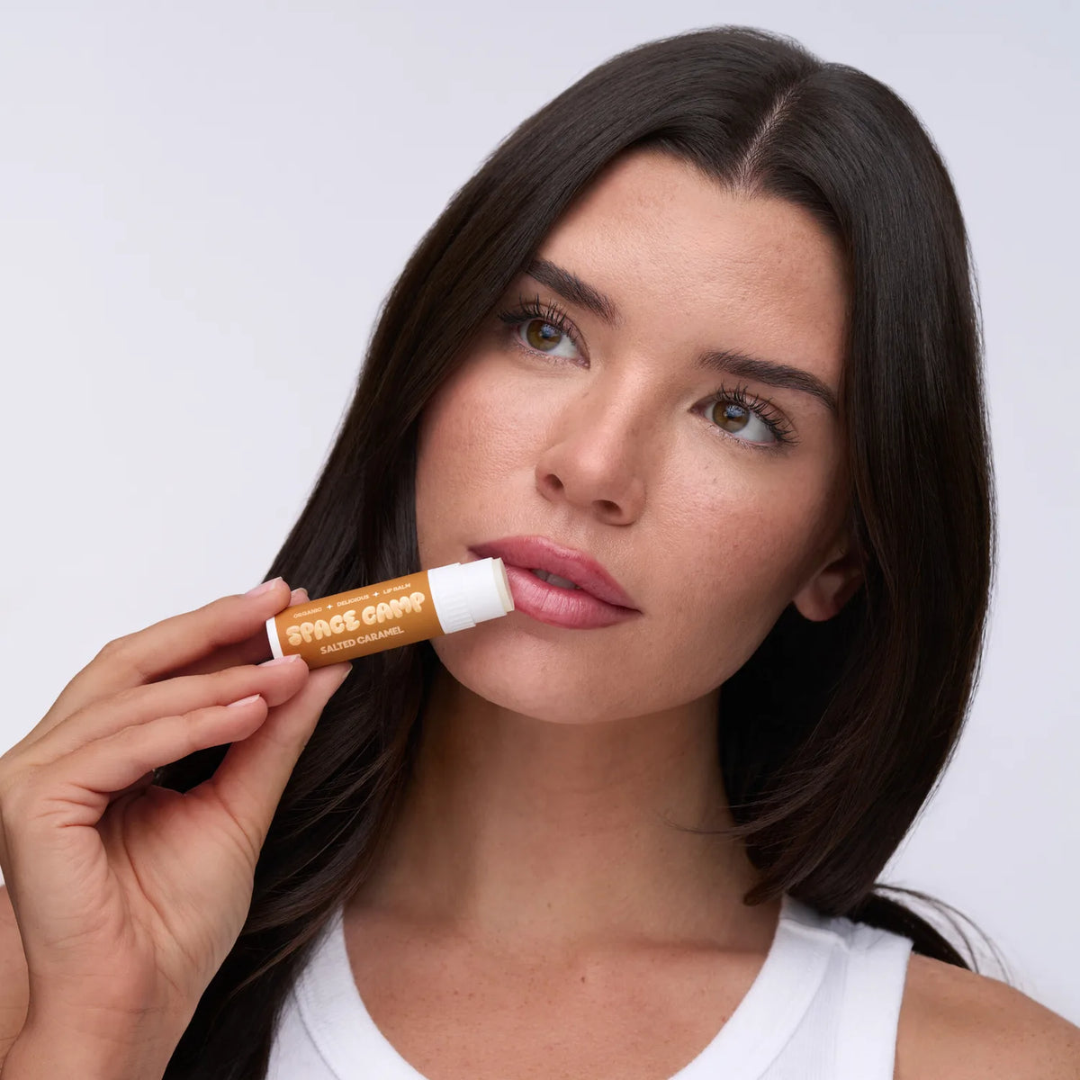 A person in a white tank top applying Space Camp Salted Caramel natural lip balm against a soft neutral backdrop.