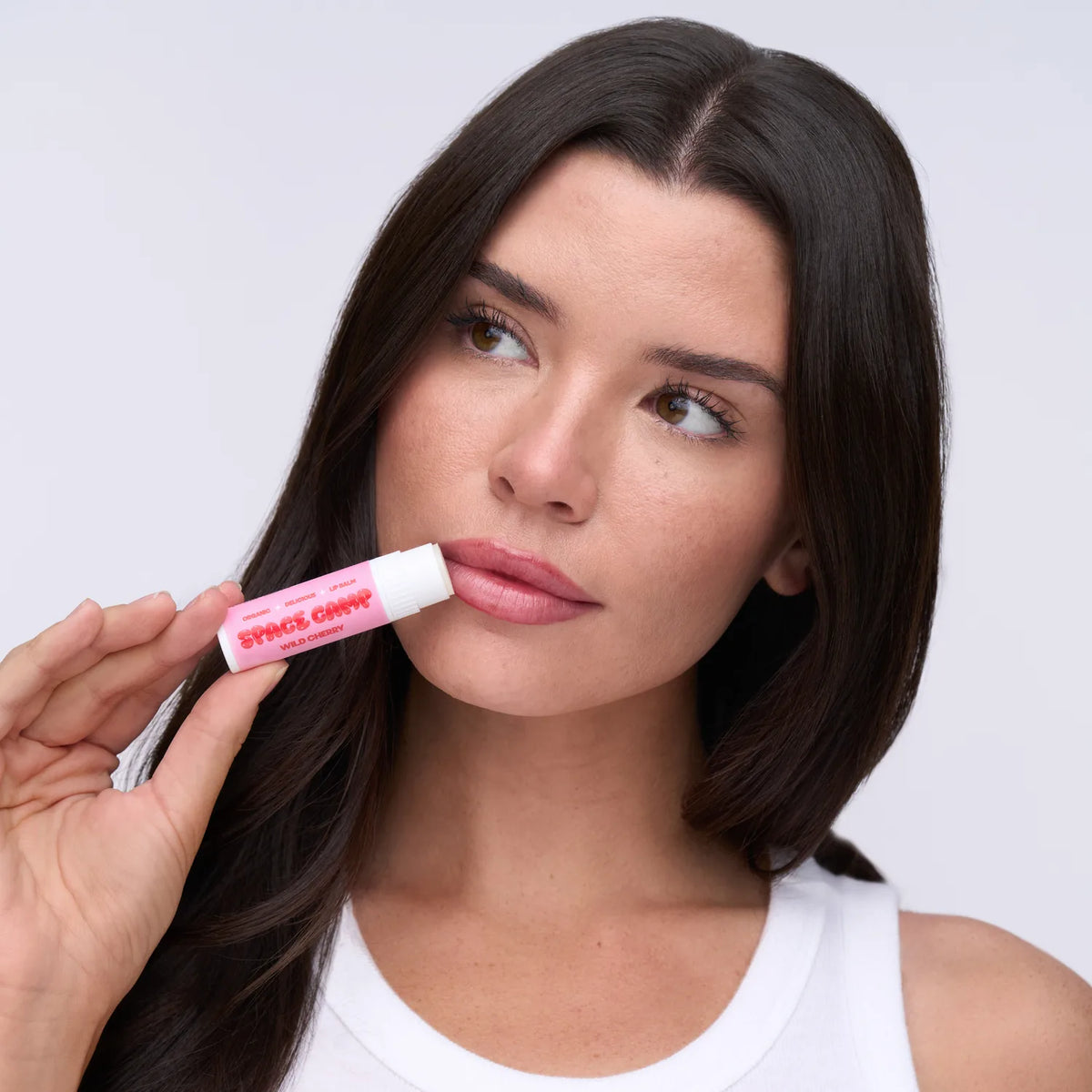 A person in a white tank top applying Space Camp Wild Cherry chapstick against a soft neutral backdrop.