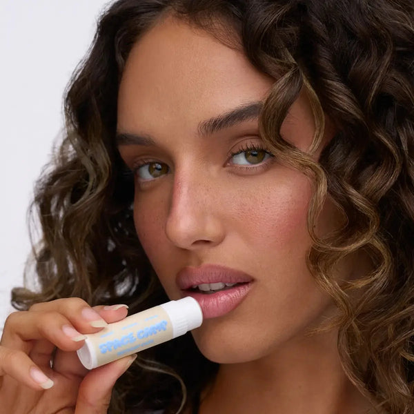 A person with curly hair applying Space Camp Birthday Cake organic chapstick in a close-up portrait.