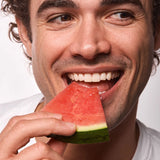 A smiling person biting into a fresh slice of watermelon, evoking the watermelon lip balm flavor.