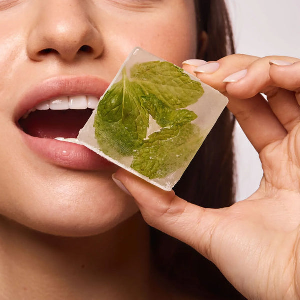 Close-up of lips holding a frosted ice cube with fresh mint leaves, evoking the cooling flavor of Space Camp Frosted Mint Lip Balm.