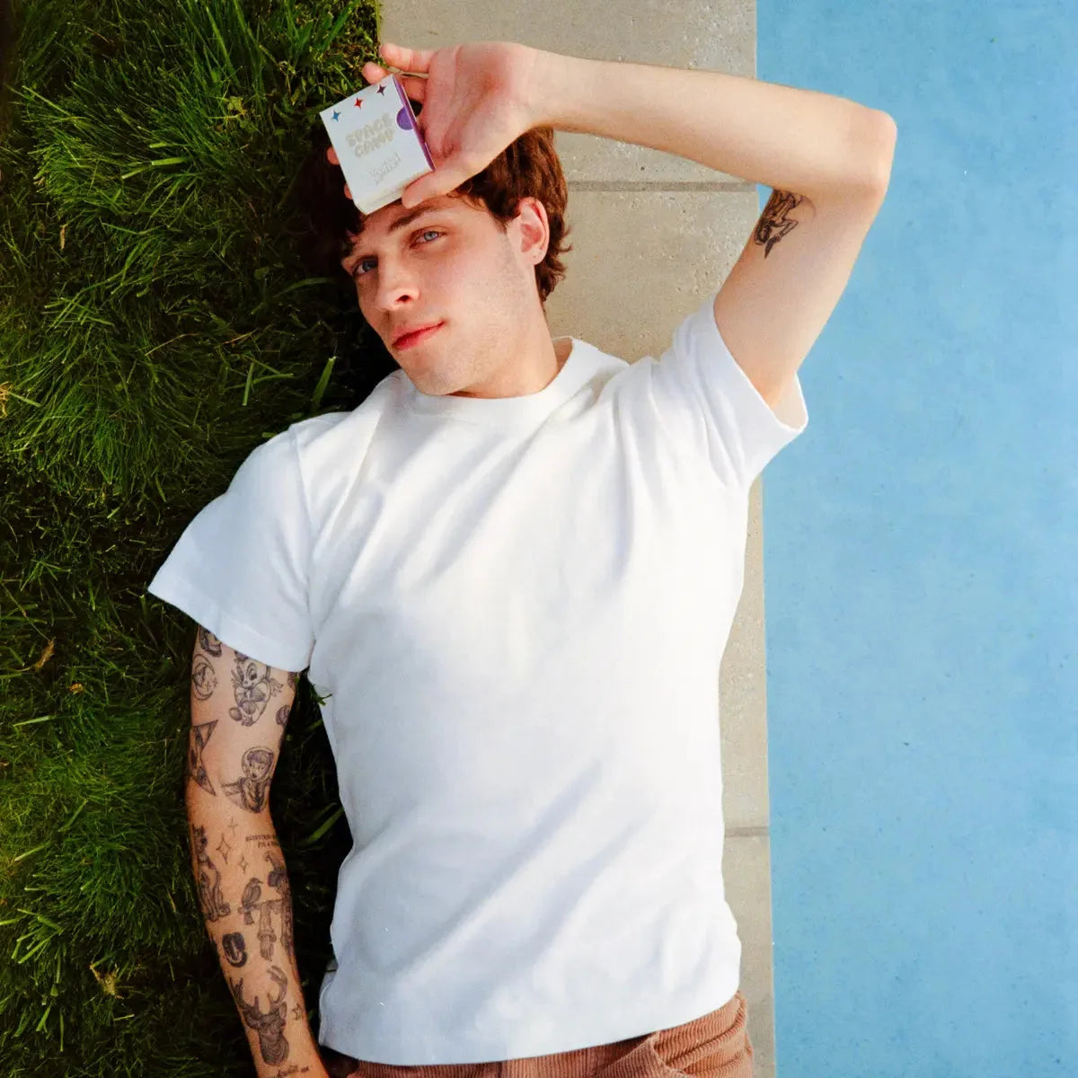 Person lying on grass by a pool holding a Space Camp Kool-Aid lip balm box.