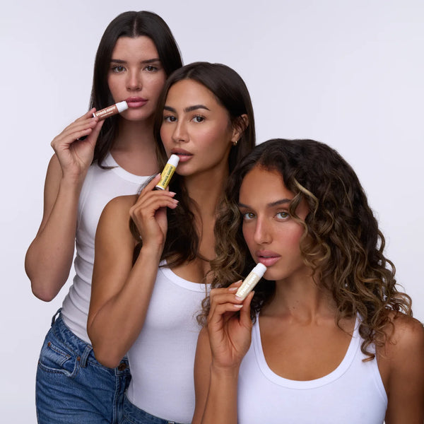 Three models applying Space Camp organic lip balm in wild cherry, tropical strawpple, and pool club pink flavors against a white background.