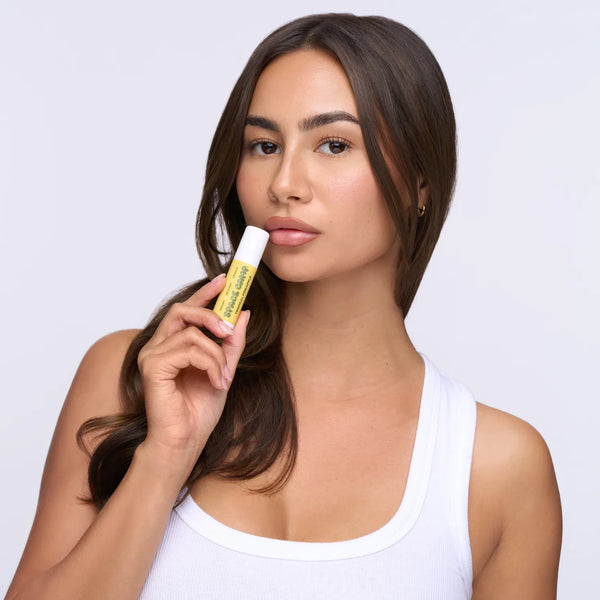 Woman with brown hair applying Space Camp Tropical Pineapple organic lip balm against a neutral background.