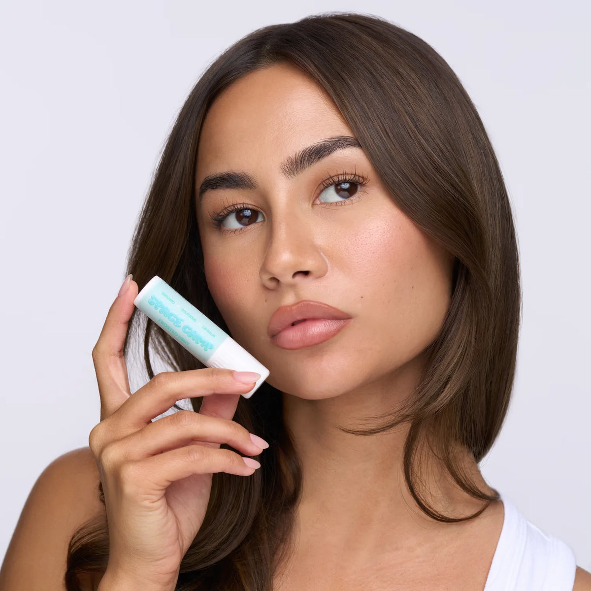 Woman with brown hair holding Space Camp Frosted Mint organic lip balm near her face against a neutral background.